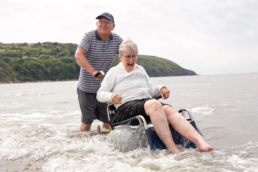 Ann laughing as she is pushed through the shallows at Poppit Sands in a wheelchair with wide balloon tyres.