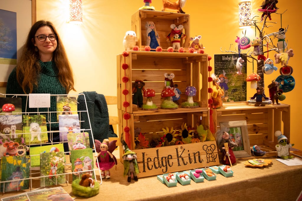 A stallholder in glasses and a green jumper beside her Hedge Kin stall of needle-felted animals, toadstools and fairies on wooden crates, next to a rack of postcards.