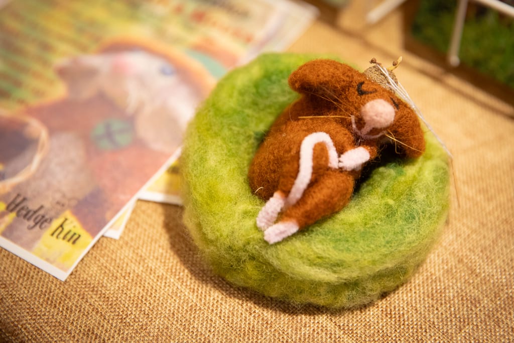 A needle-felted brown mouse curled up asleep in a green felt nest, on a hessian table beside Hedge Kin postcards.