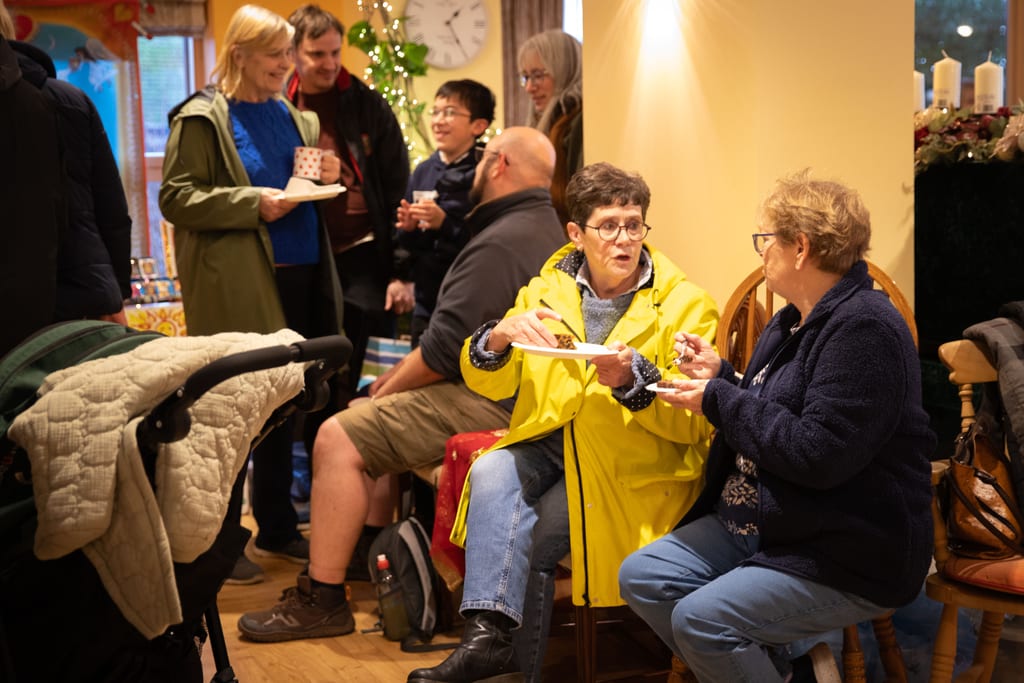 Visitors in the hospice day room with cups of tea and plates of cake, some seated and some standing, with a clock and candles behind them.