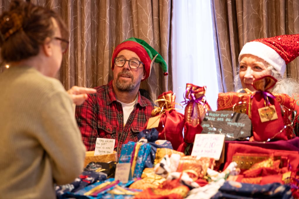 Two stallholders, one in an elf hat and one in a Santa hat, talking to a visitor across a table piled with gifts wrapped in red and gold fabric.