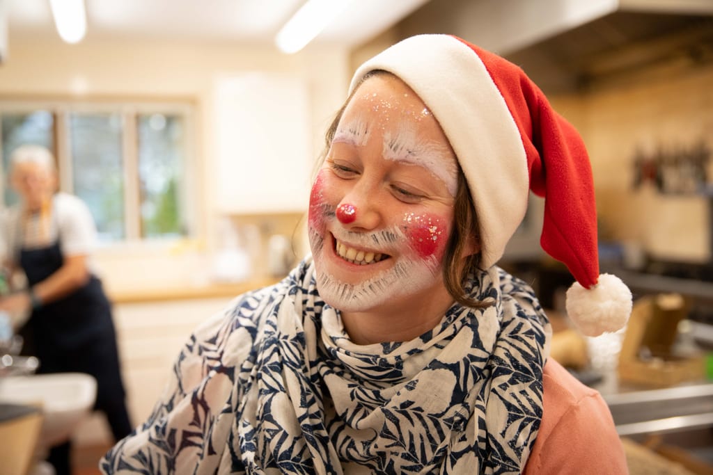A smiling woman in a Santa hat, her face painted with white eyebrows, a white beard and a red nose, in the hospice kitchen.