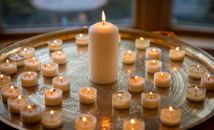 Dozens of lit tealights arranged in rings around a tall white pillar candle on a round engraved brass tray, in front of a window at dusk.