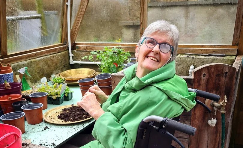 Ann, in a green coat, smiles from her wheelchair as she pots seedlings in the hospice greenhouse.