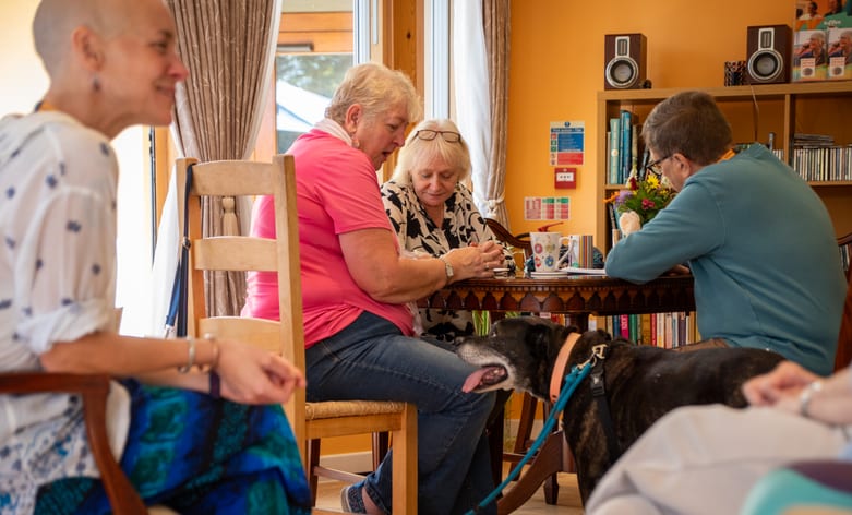 Four people sit around a small table in a bright sitting room, two of them talking over mugs of tea, with a brindle dog on a lead resting between them.