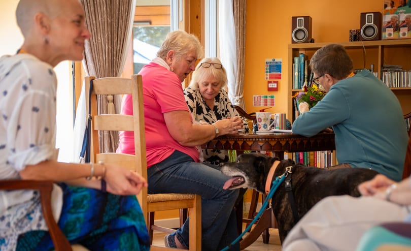 Four people sit around a small table in a bright sitting room, two of them talking over mugs of tea, with a brindle dog on a lead resting between them.