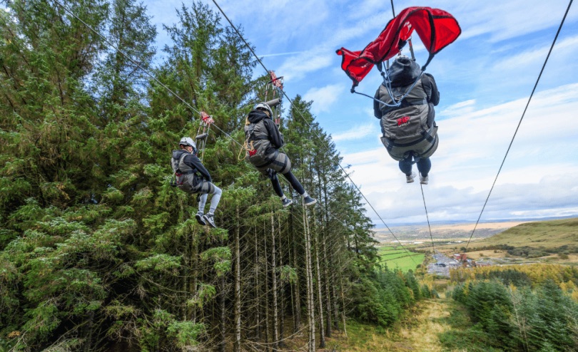 Three people harnessed to a zip wire flying above a conifer forest, a Welsh valley in the distance.